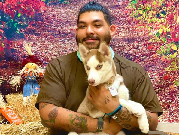 A veterinarian with tattoos on their arms holds a husky puppy with light fur in front of a colorful autumn background. There's a scarecrow decoration and straw bales to the left, enhancing the seasonal setting. Both appear content and happy.